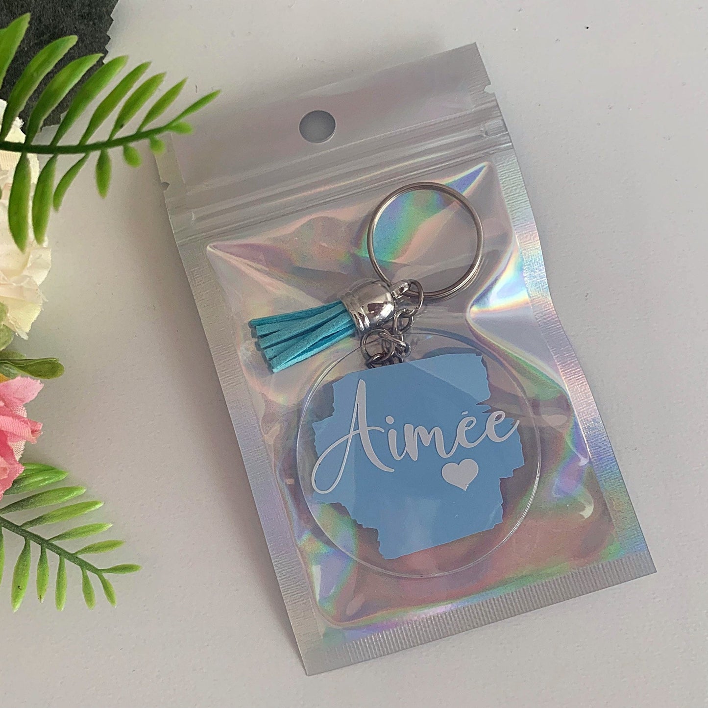 Keychain with 'Aimee' on a holographic packaging against a white background with flowers.