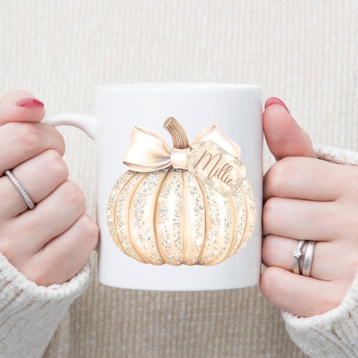 White mug with cream pumpkin design and 'Mila' branding held by two hands.