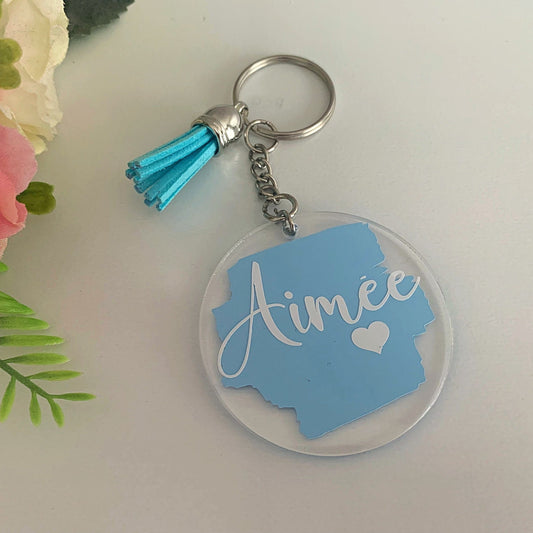 Keychain with blue state shape and 'Aimee' text, next to pink flowers on a light background