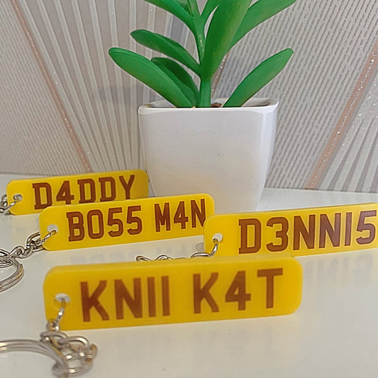 Yellow license plate keychains with text on a white surface with a plant in the background