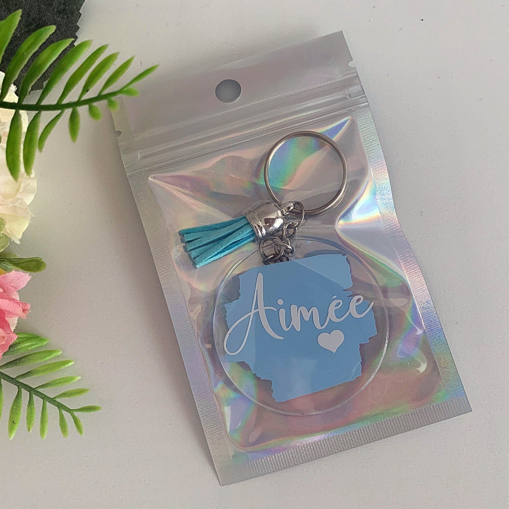 Keychain with 'Aimee' on a holographic packaging against a white background with flowers.