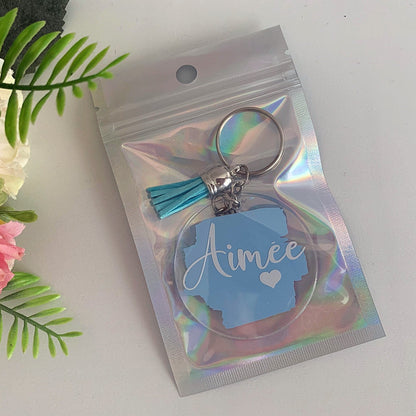 Keychain with 'Aimee' on a holographic packaging against a white background with flowers.