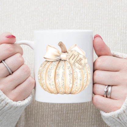 White mug with cream pumpkin design and 'Mila' branding held by two hands.