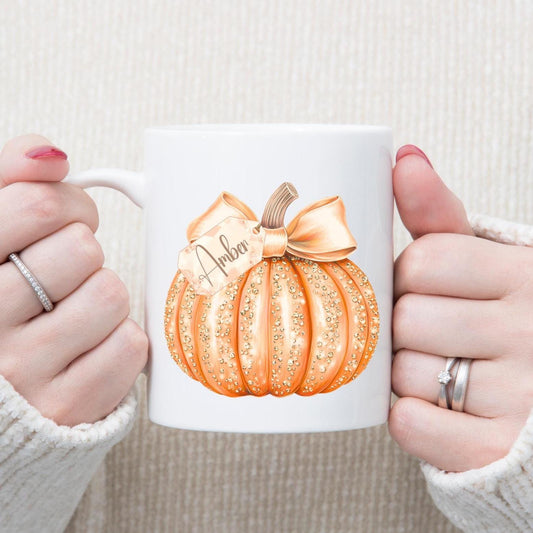 White mug with a decorative pumpkin design held by two hands against a neutral background